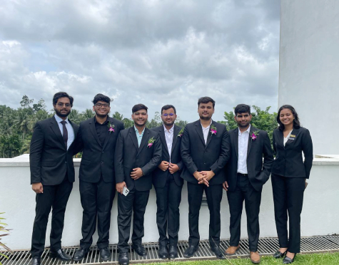 Seven people in black suits posing on a rooftop terrace; six men and one woman, all with boutonnieres, cloudy sky behind.