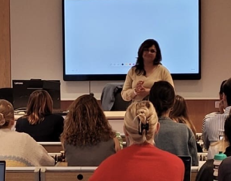 Female lecturer standing at the front of a classroom, addressing students with a large display screen behind.