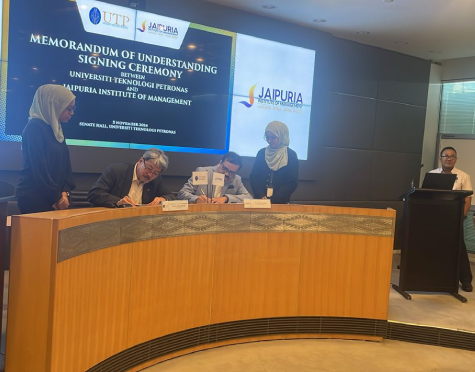 Two men sign documents at a curved desk during a memorandum of understanding ceremony, with two women standing beside and a presentation screen behind them.