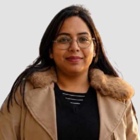 Smiling woman wearing a beige coat with faux fur collar and glasses, posing for a portrait against a light background.