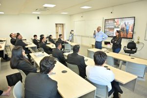 Trainer with microphone speaks to a roomful of professionals in a classroom; remote participants shown on a large monitor.