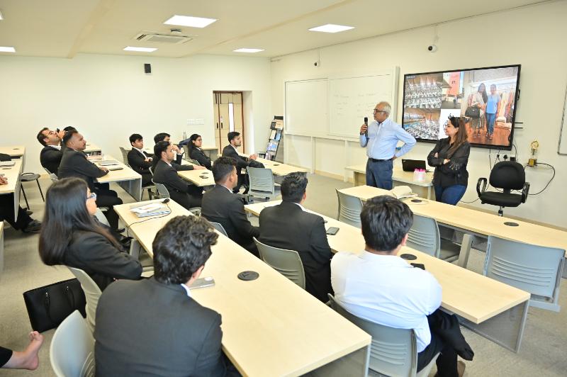 Trainer with microphone speaks to a roomful of professionals in a classroom; remote participants shown on a large monitor.